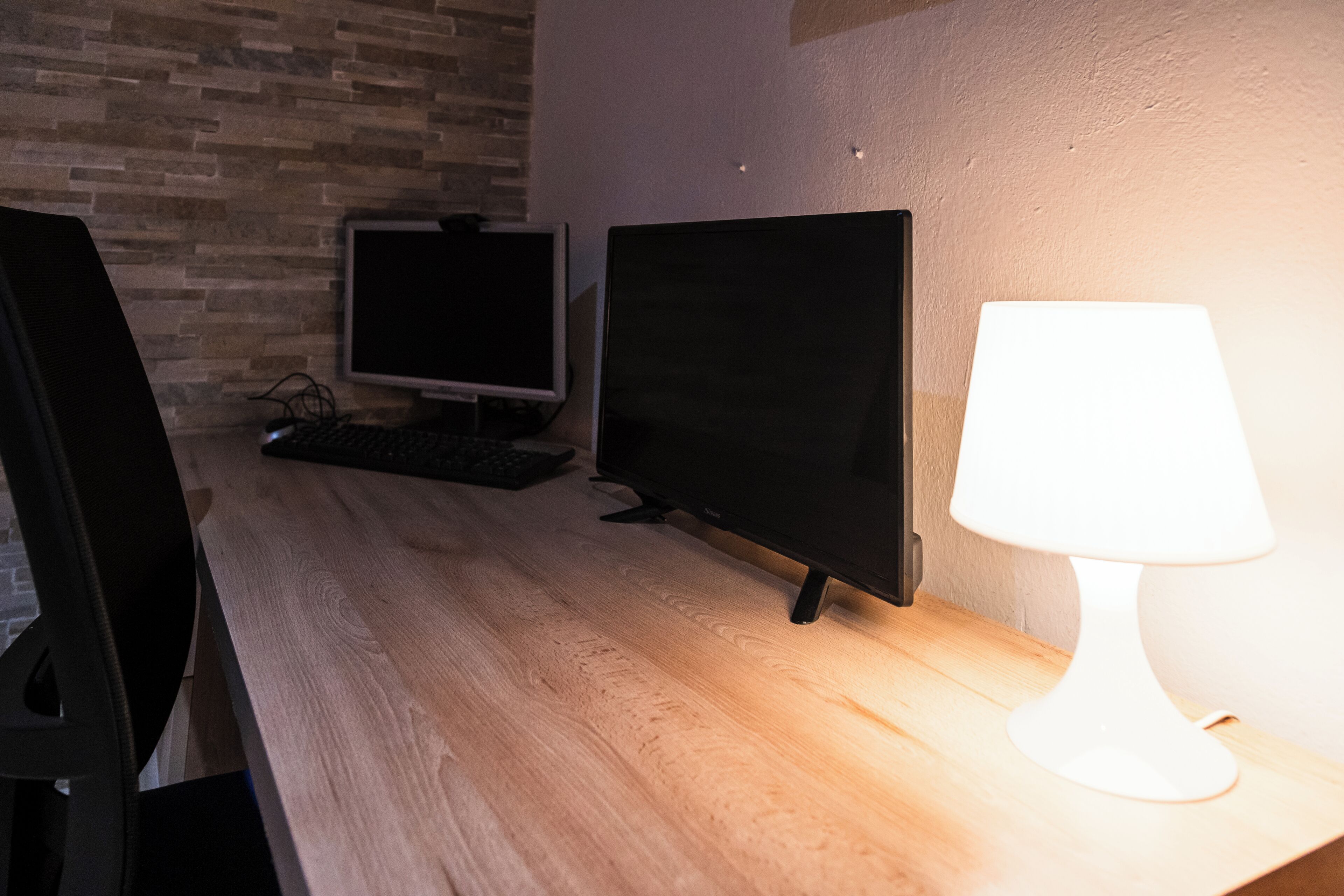 Un escritorio de madera con dos monitores, un teclado y una lámpara de escritorio blanca.