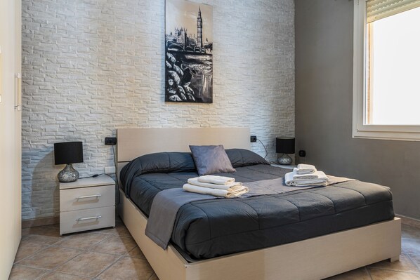 Un dormitorio moderno con pared de ladrillo, una cama grande con ropa de cama oscura, dos mesitas de noche con lámparas y una obra de arte con un paisaje urbano enmarcado sobre la cama.
