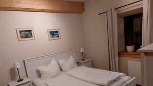 Een tweepersoonsbed met witte lakens, twee nachtkastjes met lampen, een raam met gordijnen, en twee ingelijste foto's aan de muur.