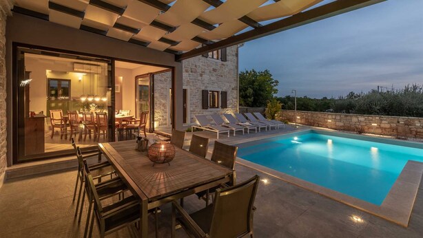 An outdoor pool area with a dining table and chairs, a stone building with a glass door, and a stone wall.