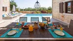 A dining table set with food and drinks, overlooking a pool with lounge chairs and a stone wall.