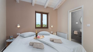 A bedroom with a bed, pillows, a bedside table with a bottle and flowers, a window with a view, and a bathroom door.
