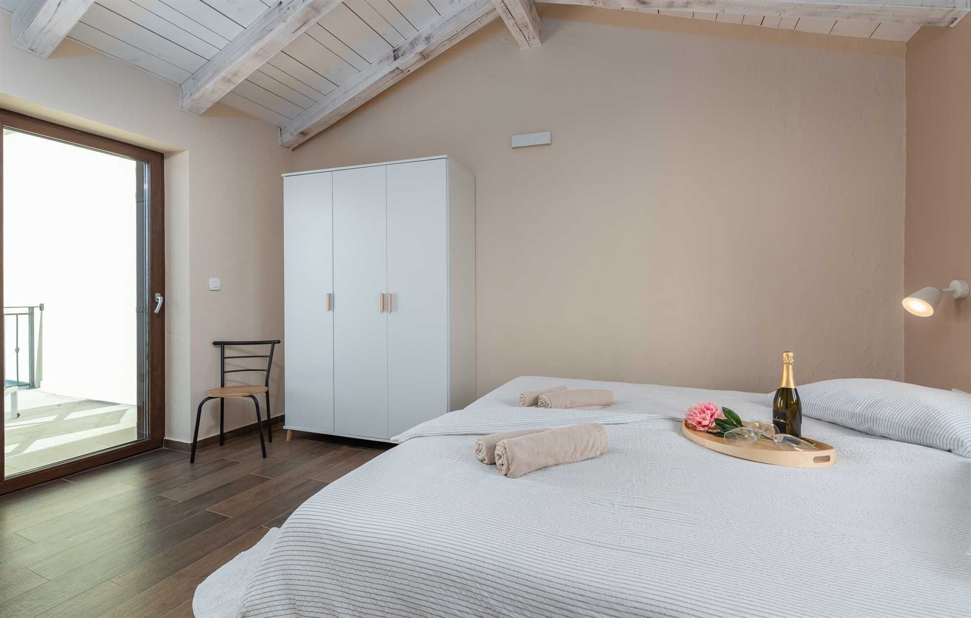 Ein Schlafzimmer mit Bett, Kleiderschrank, Stuhl und Blick auf einen Balkon, sowie eine Champagnerflasche.