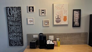 Cozinha com bancada de madeira, cafeteira, cesta de cápsulas de café e máquina de café. Há quadros nas paredes e um letreiro com a inscrição 'Coffee Mocha Latte Americano Espresso Cappuccino Macchiato Cream Sugar'.