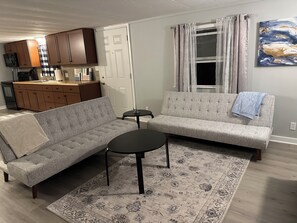 A living room with a grey sofa, a small round table, and a patterned rug. There is a kitchen in the background with wooden cabinets and a window with curtains. - "Forgey House" Just off Campus (Rio Grande)