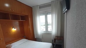 A small, neatly arranged bedroom with a single bed, a wooden wardrobe, a small table, and a TV mounted on the wall.