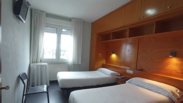 A hotel room with two single beds, a chair, a TV, and a window with curtains.