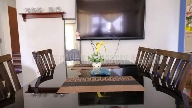 Un comedor con una mesa de vidrio, dos sillas de madera, un televisor montado en la pared y una pequeña planta en maceta sobre la mesa.