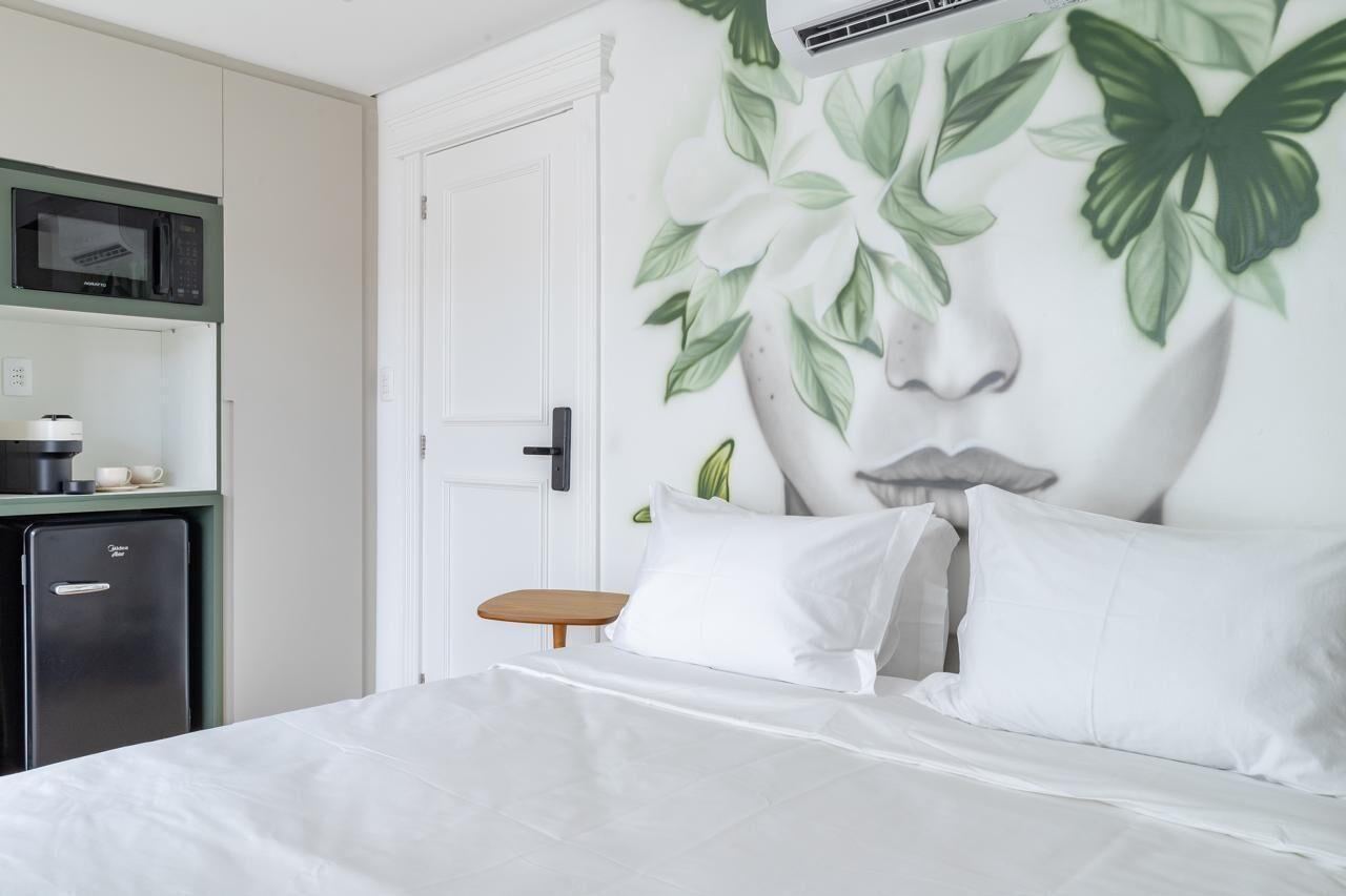 A bedroom with a bed, white bedding, a small wooden table, a microwave, and a refrigerator.