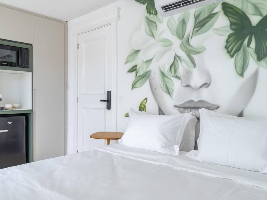 A bedroom with a bed, white bedding, a small wooden table, a microwave, and a refrigerator.
