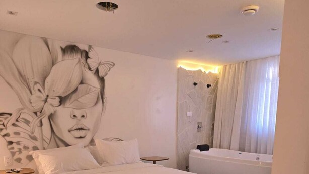 A modern bedroom with a large bed, a wall mural, a small round table, and a bathtub. - UP Hotel Boutique (Bombinhas)