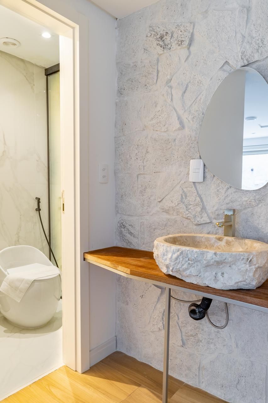 A bathroom with a stone-textured wall, a wooden vanity, a round mirror, and a white toilet.