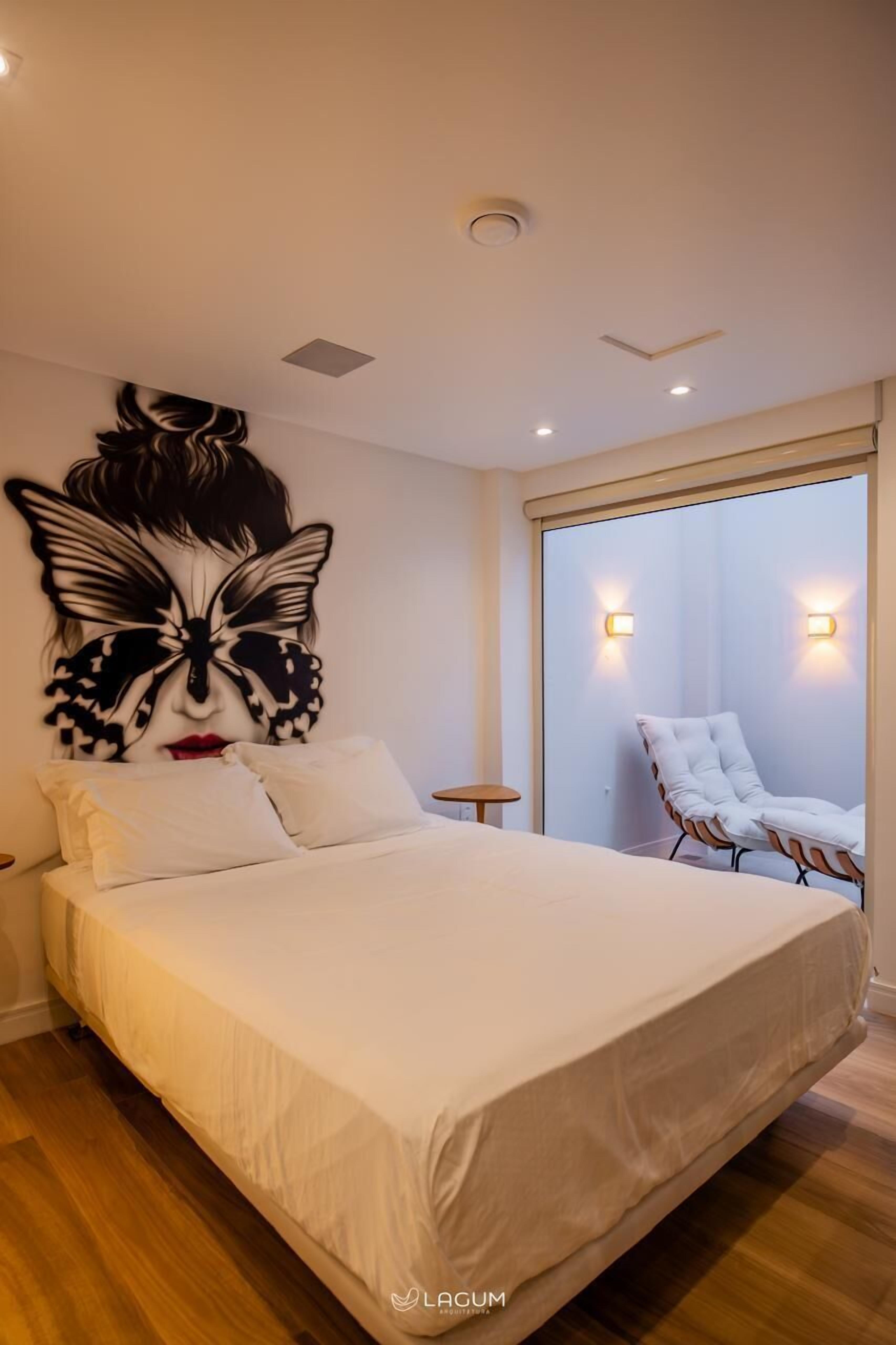 Un dormitorio con una cama grande, una obra de arte con forma de mariposa en la pared, una silla blanca y dos luces fijadas a la pared.