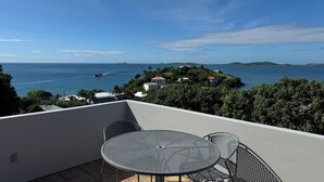 Een balkon met een tafel en stoelen, met uitzicht op de kust, een boot op het water en een klein eiland met gebouwen.