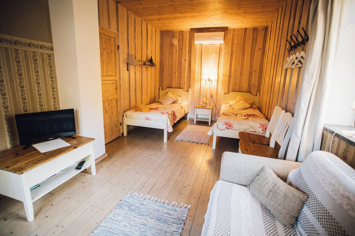 A cozy room with wooden walls, two beds with floral bedding, a white sofa, a TV on a wooden stand, and a small table.