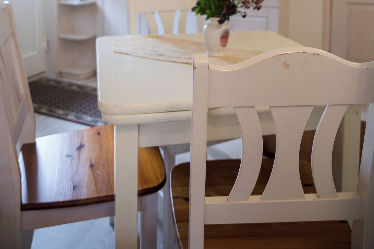 A dining area with a white table and chairs, a vase with flowers, and a kitchen in the background.