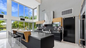 A modern kitchen with a central island, stainless steel appliances, and large windows offering a view of greenery. - Gated 9BR Estate for 20 w/ Private Pool, Jacuzzi, Theater & Guest House! Pet-Friendly! (Southwest Ranches)