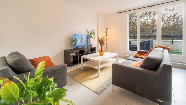 Un salon moderne avec un canapé, une table basse et un téléviseur. Une grande fenêtre offre une vue sur les arbres, et il y a un balcon.