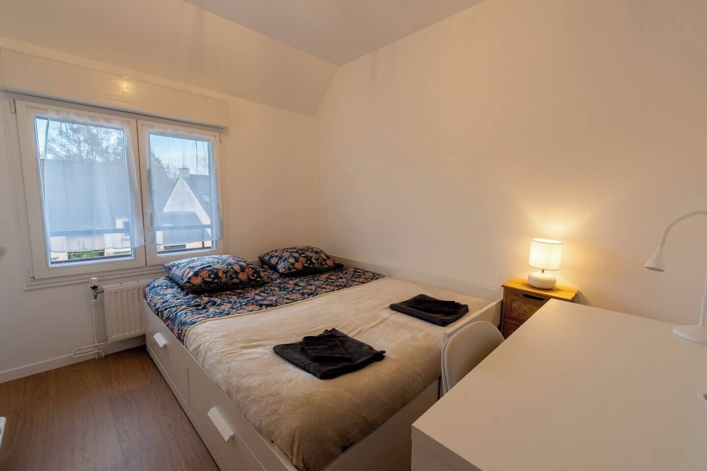 Ein kleines, ordentliches Schlafzimmer mit einem Einzelbett, einem Nachttisch mit Lampe und einem Fenster mit durchscheinenden Vorhängen.