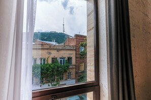 A window with a view of a building with a tower, surrounded by greenery. - Kazma Boutique Hotel (Tbilisi)