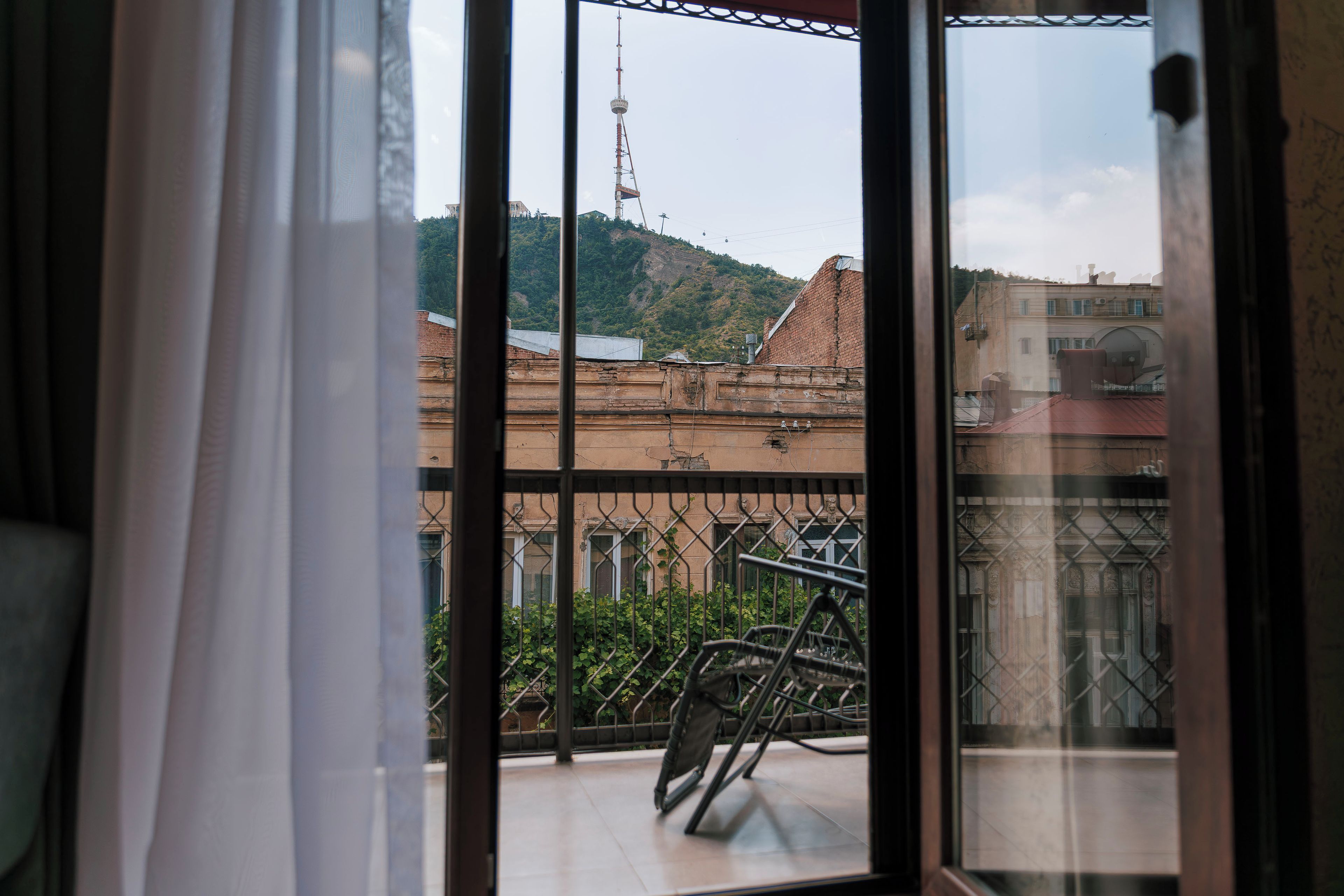 A view through a window with a metal railing, overlooking a building with a red roof and a tower in the distance.
