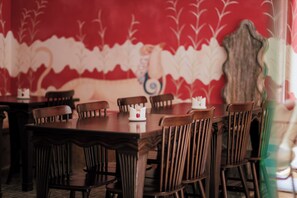Un espace repas avec des tables et des chaises en bois, un mur décoratif orné d’un motif floral et un miroir de style vintage.