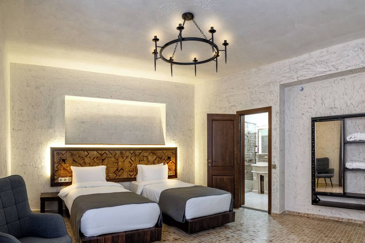 Ein Hotelzimmer mit zwei Betten, einem Holzkopfende, einer Chandelüster, einem grauen Sessel und einem großen Spiegel.
