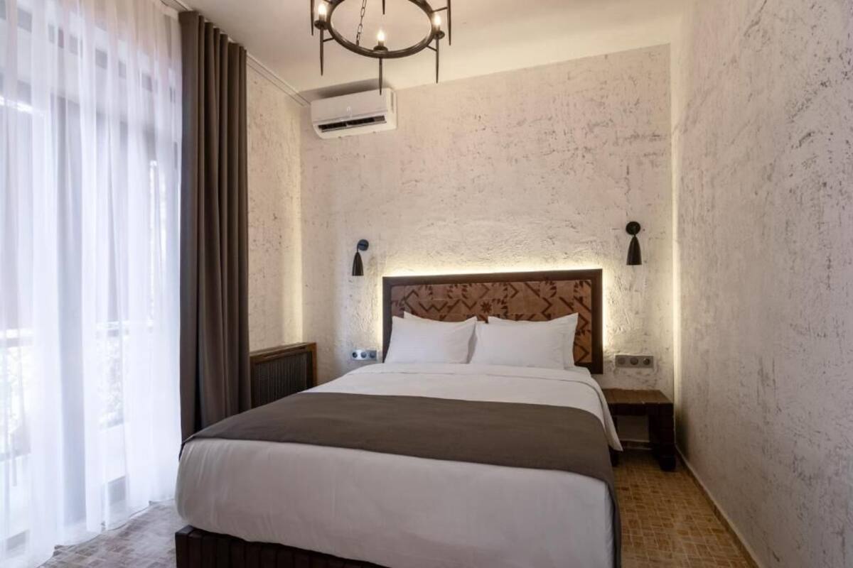 Ein Hotelzimmer mit einem großen Bett, einem Kopfende mit Muster, zwei Nachttischlampen, einem kleinen Nachttisch und einem Fenster mit leichtem Vorhang.
