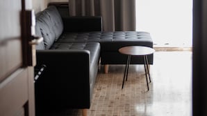 Ein modernes Wohnzimmer mit einem Ecksofa, einem kleinen runden Tisch und Blick durch Vorhänge ein Fenster.
