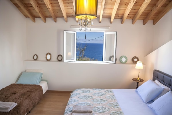 A bedroom with a large bed, a smaller bed, a window with a view of the sea, a chandelier, and wall decorations.