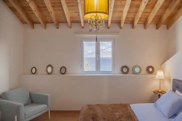 A bedroom with a wooden ceiling, a large window with a view, a bed with a fluffy blanket, a chair, and a bedside table with a lamp. - Water from Alto Main House (Água de Alto)