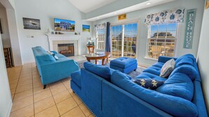 Een woonkamer met een open haard, blauwe banken, een plafondventilator en uitzicht op het strand.
