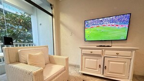 Ein Wohnzimmer mit einem Rattan-Sofa, einem weißen Schrank und einem Fernseher, der ein Fußballspiel zeigt.