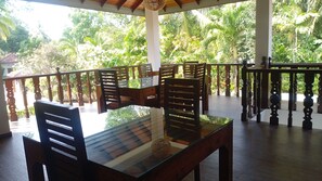 A covered outdoor seating area with wooden furniture and a glass table, surrounded by a wooden railing with a view of lush greenery. - Rockery Villa Ayurveda (Bentota)