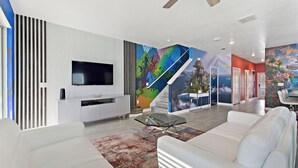 A modern living room with a flat-screen TV, white sofas, a glass coffee table, and a colorful mural on the wall. - Wonderland Sleeps 14 Free Waterparks and South Facing Pool (Kissimmee)