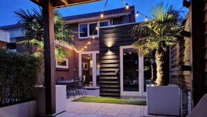 Une terrasse avec une table en bois et deux chaises rembourrées, une maison en briques dotée d’une porte-fenêtre coulissante et des guirlandes lumineuses.