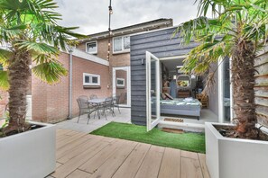 Eine moderne Terrasse mit Holzboden, einem kleinen Tisch und Stühlen sowie einem Bett mit Blick durch eine offene Tür.