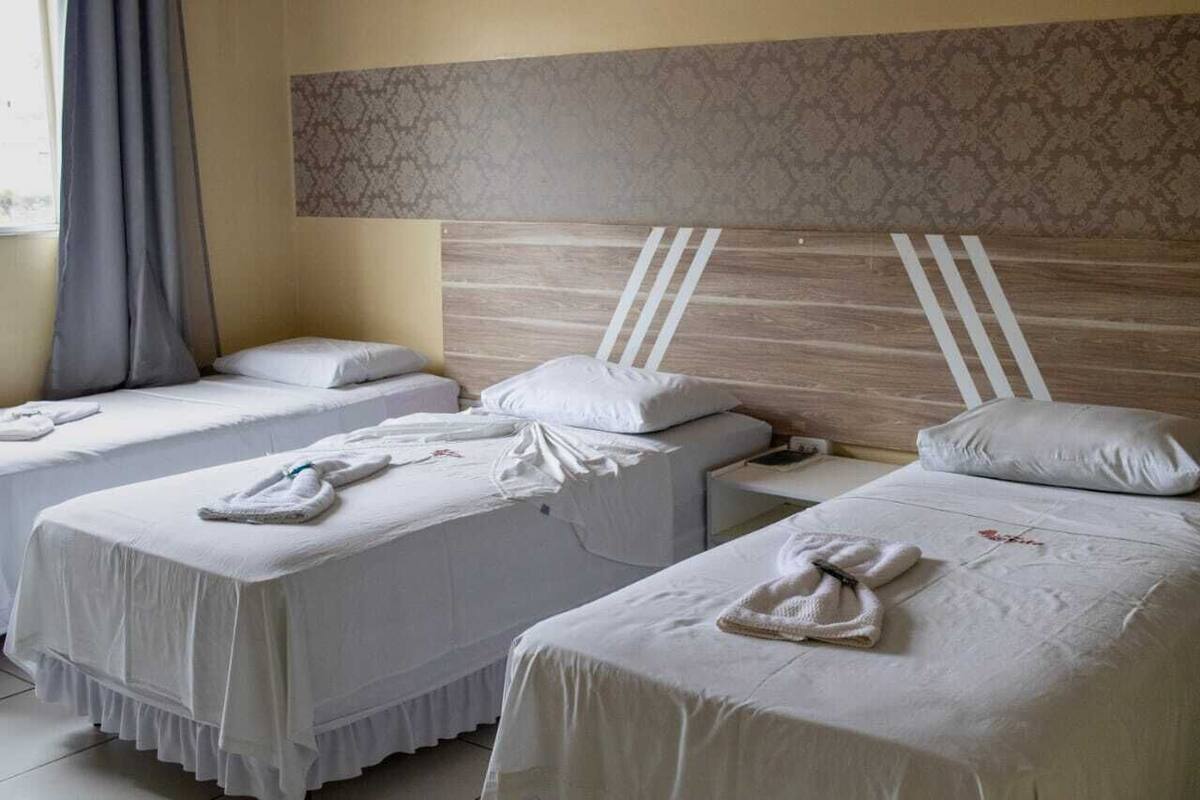 A hotel room with three single beds, each with a white bedspread and pillows, and a headboard with a wooden panel and white stripes.