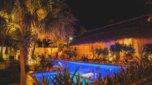 A nighttime pool area with a thatched-roof building, surrounded by tropical plants. - Bidmont Villa Spa (Puerto Escondido)