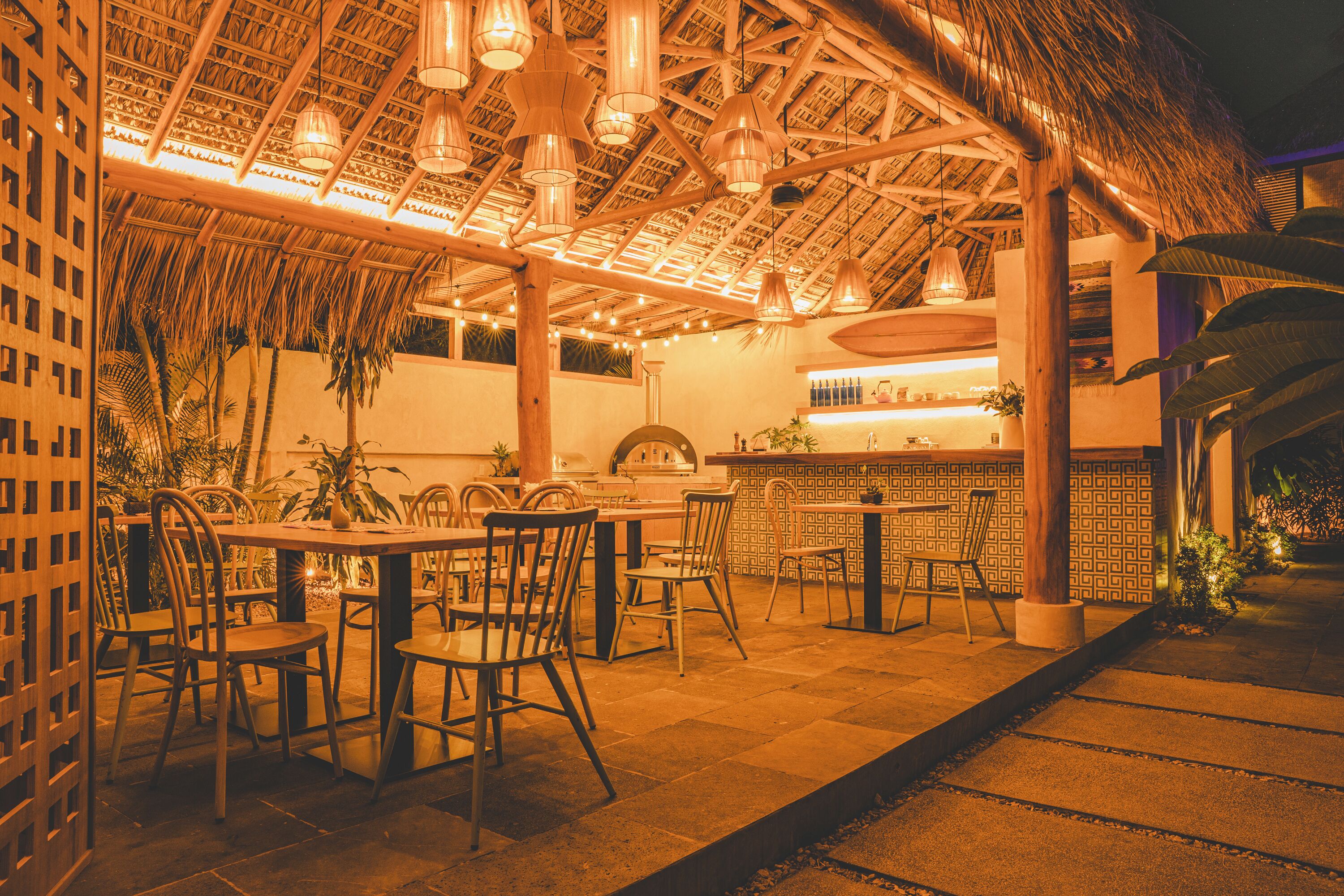 Un área de comedor al aire libre con mesas y sillas de madera, techo de paja y lámparas colgantes.