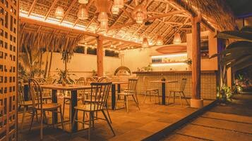 Un área de comedor al aire libre con mesas y sillas de madera, techo de paja y lámparas colgantes.