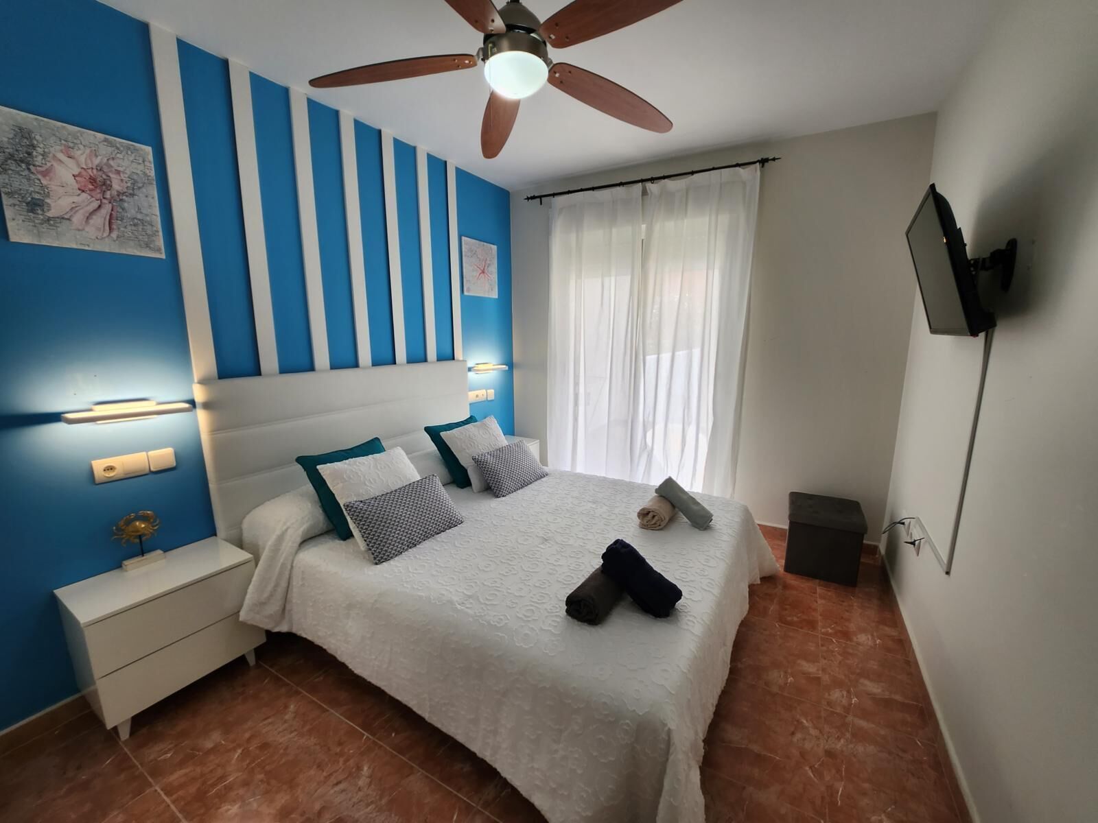 A bedroom with a bed, a nightstand, a ceiling fan, a TV, and a window with curtains.