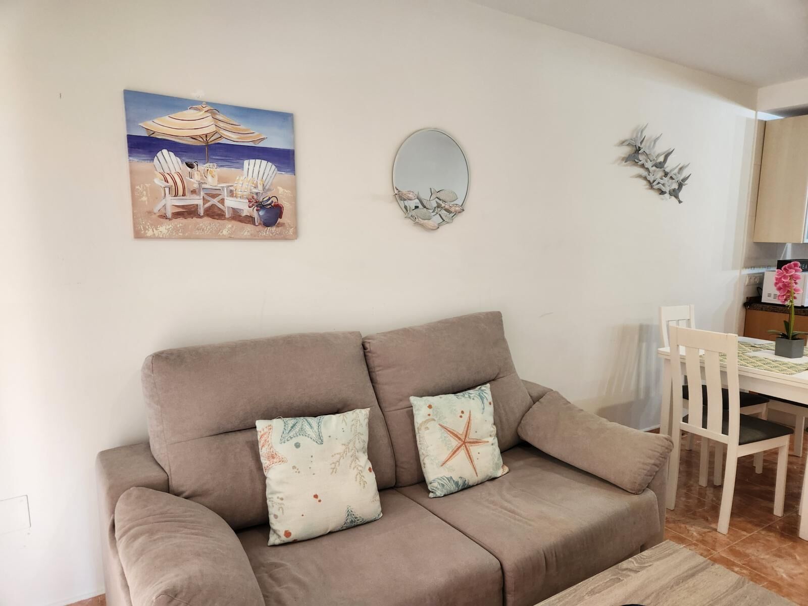 Een woonkamer met een beige bank, twee decoratieve kussens, een schilderij van een strandtafereel, een ronde spiegel en een aan de muur bevestigde plant.