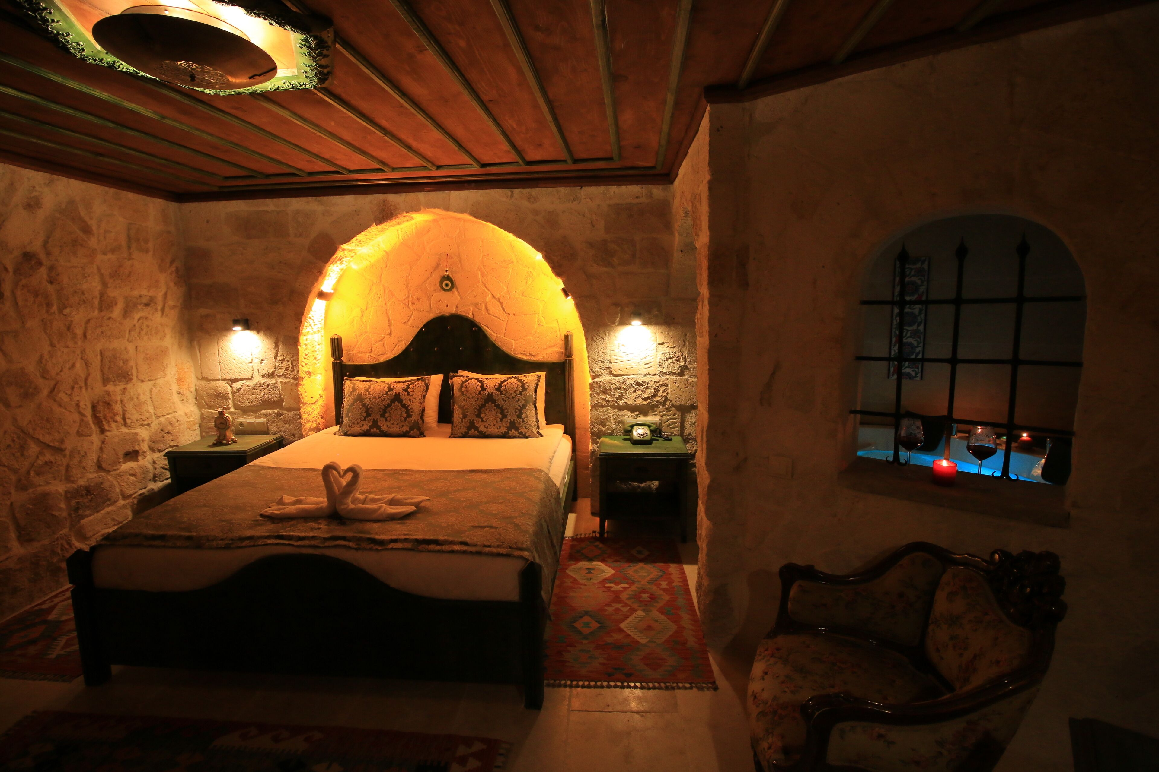 A cozy bedroom with a stone wall, a wooden ceiling, a bed with a heart-shaped pillow, a nightstand, a chair, and a window with a view.