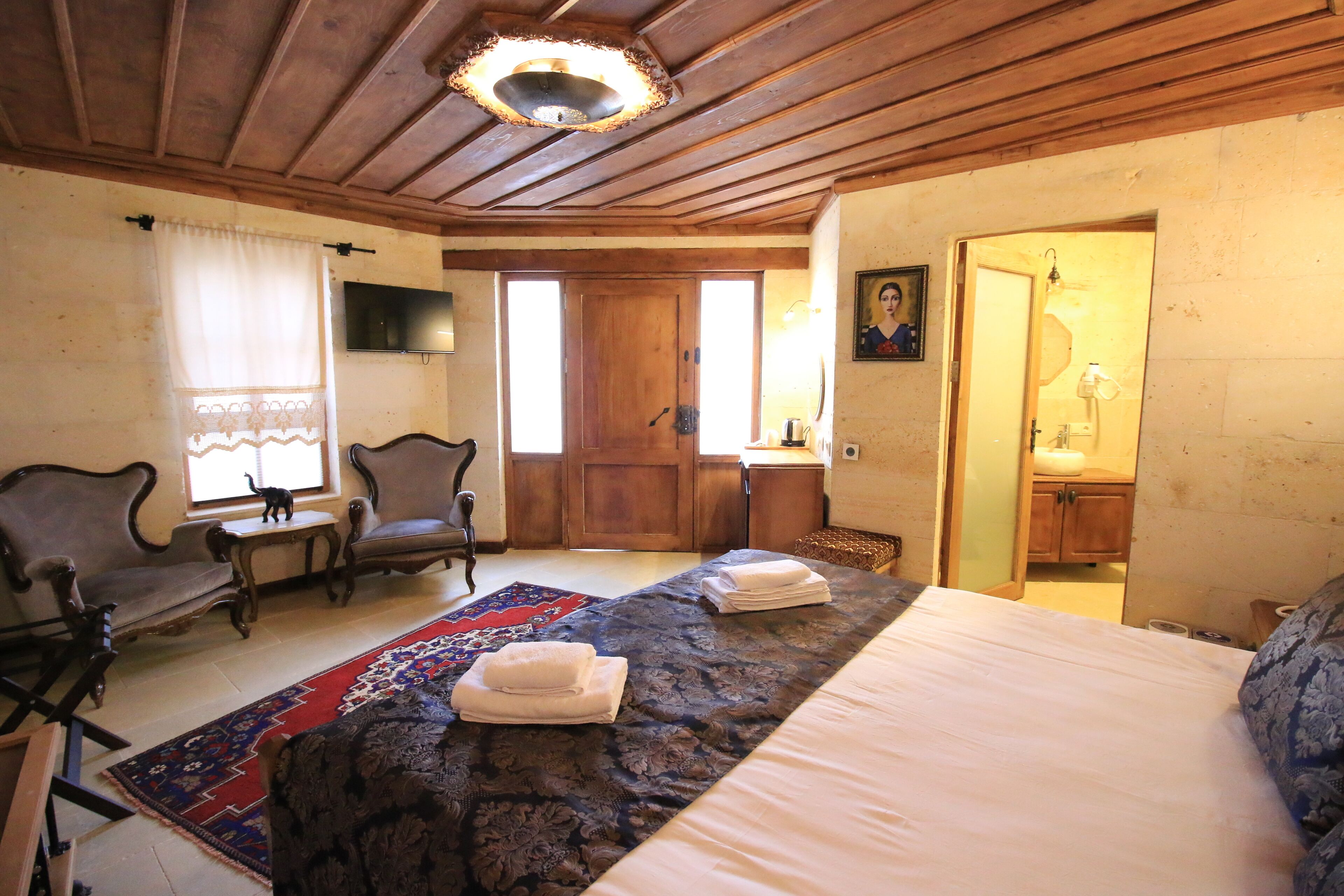 Una habitación con una cama, dos sillones, techo de madera, televisor y una puerta que da a un baño.
