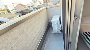 Un balcón estrecho con piso de madera, un equipo de aire acondicionado blanco y vista a edificios vecinos.