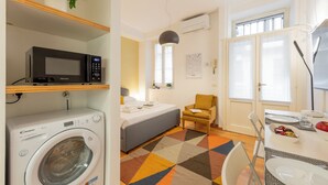 Ein kleines Hotelzimmer mit Waschmaschine, einem Bett, einer Couch, einem Esstisch mit Stühlen und einem fenster mit Vorhängen.