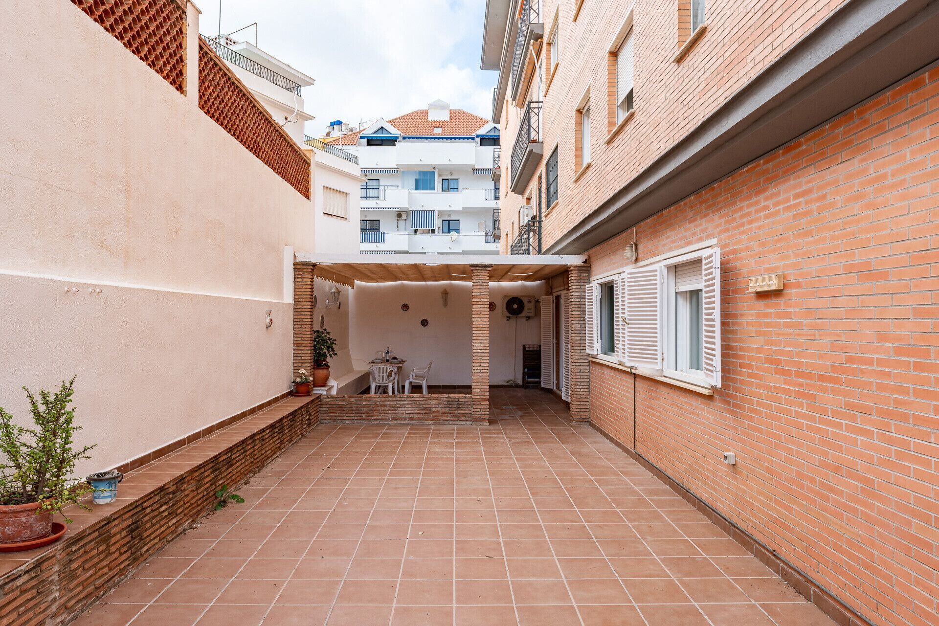 A narrow courtyard with tiled flooring, a white table and chairs, and a potted plant.