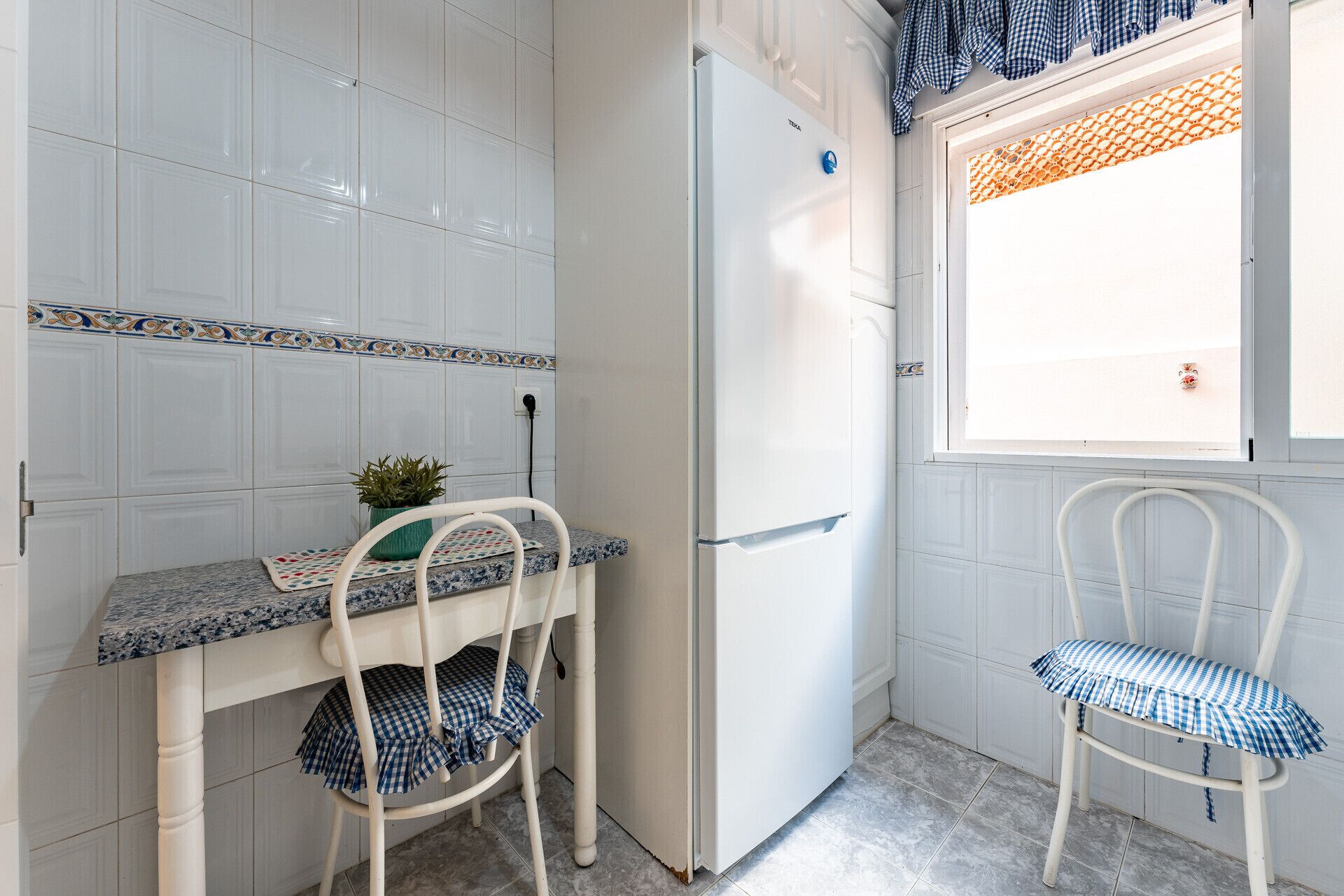 A compact kitchen with white tiled walls, a white refrigerator, a small table with a plant, and two chairs.