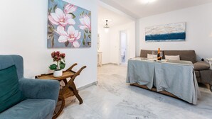 A living room with a sofa, a dining table with a bottle and glasses, a painting of flowers, and a view into another room. - MalagaSuite Terrace&Parking Stay Fuengirola (Fuengirola)